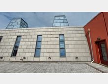 Moderne Gebäudefassade mit beiger Verkleidung aus rechteckigen Platten und schmalen, vertikalen Fenstern; zwei gläserne pyramidenförmige Dächer ragen heraus. Rechts ist eine rote Wand mit einer Tür und einer Regenrinne sichtbar.