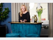 Eine Frau steht hinter einer Bar mit einer blau gepolsterten Theke und goldenem Rand. Auf der Theke stehen ein Blumenstrauß in einer Vase, ein goldener Behälter in Ananasform und ein Getränkeshaker, im Hintergrund sind beige Vorhänge vor einem Fenster sichtbar.