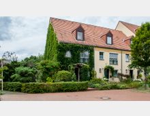 Reihenhaus mit hellgelber Fassade und rotem Ziegeldach, dessen linke Seite vollständig mit grünem Efeu bewachsen ist. Davor ein kleiner Vorgarten mit Blumen, Sträuchern und einem gepflasterten Gehweg; Straßenschild mit der Aufschrift „Himbeersteig“ am linken Bildrand.