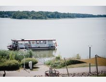 Ein kleiner Dampfer mit der Aufschrift "Mein Dampfer" liegt an einem Holzsteg in einem See, umgeben von grüner Vegetation. Im Hintergrund ist eine bewaldete Uferlandschaft zu sehen, während im Vordergrund ein gepflasterter Weg und eine Sonnenschutzkonstruktion sichtbar sind.