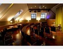 Dachgeschoss-Veranstaltungsraum mit schrägen Decken, Holzfußboden und bogenförmig angeordneten Stuhlreihen vor einer kleinen Bühnenfläche mit gelbem Vorhang. Entlang der Wand stehen gepolsterte Sitzbänke mit kleinen runden Tischen und Tischlampen; an der Decke hängen Scheinwerfer und eine Diskokugel, hinten sind zwei Fenster.