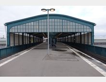 Bahnhof mit überdachtem Bahnsteig, bestehend aus einer grünen Stahlkonstruktion mit großen Glasflächen. Im Zentrum des Bildes befindet sich eine Sitzbank, vereinzelt sind Personen auf dem Bahnsteig zu sehen.