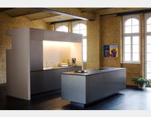 Moderne Küche in einem Raum mit gelben Ziegelwänden und großen Fenstern mit Rundbögen. Die Küche besteht aus einer grauen Wandzeile mit eingelassener Beleuchtung und einer freistehenden Kücheninsel, ergänzt durch eine schlichte Einrichtung und Zimmerpflanzen.