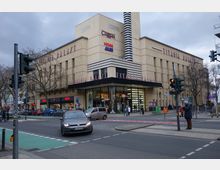 Straßenszene mit dem „Titania Palast“, einem großen Gebäude mit cremefarbener Fassade, breiter Fensterfront und vertikaler Streifenstruktur. Im Erdgeschoss befinden sich Geschäfte wie HEMA, während vor dem Gebäude Autos, eine Ampel und Fußgänger zu sehen sind.