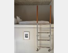 Loft-style sleeping area with a low concrete ceiling, featuring a bed with white bedding and a wall-mounted metal ladder with wooden handrails leading up to it. A plain white wall below includes a small framed artwork.