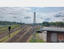 Bahngelände mit mehreren Gleisen, Oberleitungen und vereinzelten Signalanlagen, umgeben von dichter Vegetation im Hintergrund. Rechts im Bild steht ein Backsteingebäude mit flachem Dach, daneben ein Parkplatz mit geparkten Autos.