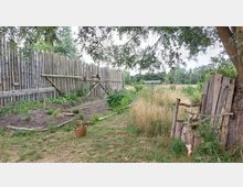 Ein naturnaher Garten mit einem hölzernen Zaun im Hintergrund, einem kleinen bepflanzten Beet und wild wachsendem Gras. Rechts steht ein einfacher Holzstuhl neben einem Baum, daneben liegt ein Weidenkorb.