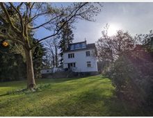 Freistehendes, weißes Wohnhaus mit mehreren Stockwerken und Dachgauben, über eine breite Treppe von einem großen, grünen Garten aus erreichbar. Umgeben von hohen Bäumen und Sträuchern unter hellem Himmel mit tief stehender Sonne.