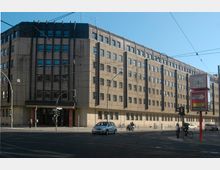 Ein großes, rechteckiges Bürogebäude aus Beton mit vielen gleichmäßig angeordneten Fenstern und einem zentralen Eingang; im Vordergrund eine Straßenkreuzung mit Ampeln, Fußgängern, Autos und Fahrrädern.