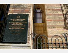 Außenansicht einer Synagoge mit hellbraun-rot gemusterten Backsteinwänden. Im Vordergrund ist eine schwarze Gedenktafel mit goldener Schrift angebracht, daneben ein verziertes Fenster und ein dekoratives Metallgitter.