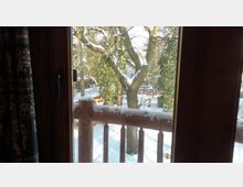 Blick durch ein Fenster auf einen verschneiten Garten mit Bäumen, Zaun und einem Haus im Hintergrund. Ein Holzgeländer im Vordergrund ist ebenfalls mit Schnee bedeckt.
