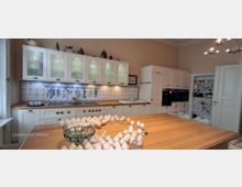 Große, helle Wohnküche mit weißen Einbauschränken und Milchglas-Oberschränken, Holzarbeitsplatte und blau-weiß gefliestem Fliesenspiegel mit dekorativen Motiven. Im Vordergrund steht eine große Kücheninsel aus Holz; rechts sind Hochschränke mit Einbauöfen und ein offenes Regal neben einer weißen Tür.