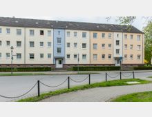 Mehrfamilienhaus mit drei Stockwerken und einem Dach mit dunklen Ziegeln. Die Fassade ist in Beige- und Blautönen gestrichen, umgeben von Hecken, Gehweg und einer Straße im Vordergrund.