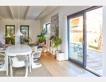 Helles Wohnzimmer mit modernem Essbereich, weißen Möbeln und großen Fenstern, die auf eine Terrasse mit Holzboden führen. Zimmerpflanzen und Regalelemente prägen die Einrichtung, während der Holzboden und die Lichtgestaltung eine warme Atmosphäre schaffen.