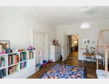 Kinderzimmer mit heller Einrichtung und Holzfußboden. Links eine weiße Regalwand mit Büchern und Spielzeug, rechts ein kleiner Schreibtischbereich, in der Mitte ein bunter Teppich mit Blumenmuster; im Hintergrund eine geöffnete Tür mit Blick in einen weiteren Raum.