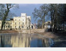 Gelbes Schloss im neogotischen Stil mit Zinnen und großen Fenstern, gelegen an einem Teich mit Spiegelung des Gebäudes im Wasser; umgeben von Bäumen und weiteren Gebäuden, darunter ein kleiner Pavillon.