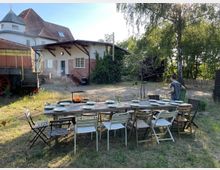Ein weitläufiger Garten mit einem langen, gedeckten Holztisch und Stühlen im Vordergrund, daneben lodert ein kleines offenes Feuer. Im Hintergrund stehen ein altes Ziegelhaus mit Satteldach und ein Eisenbahnwaggon, umgeben von Bäumen und Wiese.