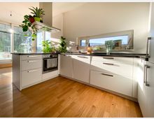 Moderne Küche mit weißen Schränken in L-Form, einer schwarzen Arbeitsplatte und integriertem Backofen. Große Fenster lassen viel Tageslicht herein, und mehrere Zimmerpflanzen sind auf der Arbeitsfläche dekorativ platziert.