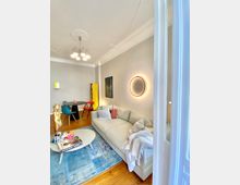 Wohnzimmer mit modernem Design, bestehend aus einer hellen Sofalandschaft, rundem Couchtisch und einem blauen Teppich auf Holzfußboden. Im Hintergrund befindet sich ein Esstisch mit bunten Stühlen, gelber Regalwand und minimalistischer Beleuchtung; an den Wänden hängen Bilder und eine runde leuchtende Wandlampe.