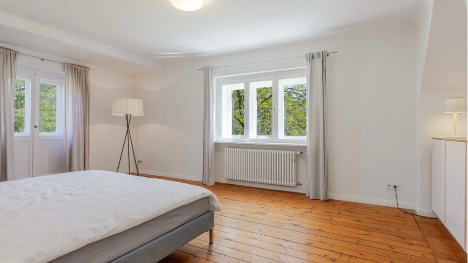 Helles Schlafzimmer mit weiß gestrichenen Wänden, Holzdielenboden und großem Fenster mit Vorhängen sowie Heizkörper darunter. Im Raum stehen ein Bett, eine Stehlampe und ein Sideboard; links ist eine verglaste Tür mit Vorhängen sichtbar.