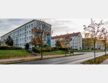 Wohngebiet mit mehrstöckigen Wohnhäusern aus hell verputztem Beton, umgeben von kleinen Grünflächen und Herbstbäumen. Eine Straße mit Gehwegen verläuft durch die Szene, im Vordergrund sind fallende Blätter zu sehen.