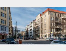 Straßenszene mit mehrstöckigen Wohngebäuden im Altbaustil, beidseitig der Straße. Im Vordergrund stehen parkende Autos und Straßenschilder, darunter ein Schild mit der Aufschrift „Eisenacher Str.“, sowie vereinzelte Bäume und Geschäfte im Erdgeschoss.