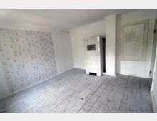 Ein leerer Raum mit weiß gestrichenen Wänden, teils mit gemustertem Tapetenabschnitt in Blau und Beige. Der Boden besteht aus abgenutzten Holzdielen, und in einer Ecke steht ein weiß gekachelter Ofen.