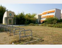Eingangsbereich einer S-Bahn-Station mit einem markanten "S"-Symbol auf einem beige-farbenen Gebäude. Im Vordergrund befinden sich Fahrradständer, und im Hintergrund sind Bäume sowie ein modernes Gebäude mit orangefarbenen Markisen zu sehen.