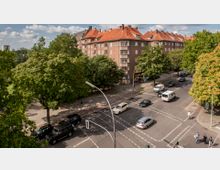 Straßenkreuzung in einem urbanen Wohngebiet mit mehrstöckigen, roten Ziegelhäusern und roten Dächern, umgeben von dichtem Baumbestand. Auf der Straße fahren mehrere Autos, während Fußgänger und Radfahrer den Bereich queren.