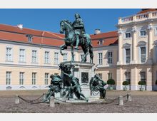 Reiterstandbild aus Bronze auf einem detailliert verzierten Sockel, umgeben von vier sitzenden Figuren, in einem gepflasterten Hof vor einer gelben Barockfassade mit weißen Fenstern und roten Ziegeldächern. Himmel ist klar und blau.