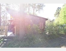 Holzhütte in einem bewaldeten Gebiet, mit einer Veranda und einer angrenzenden umzäunten Fläche. Umgeben von Bäumen und Sträuchern, führt ein Kiesweg an der Hütte vorbei, während Sonnenlicht durch die Baumkronen scheint.