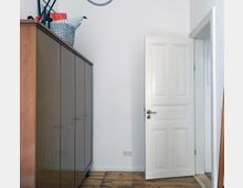 Ein schlichter Innenraum mit einem hölzernen Kleiderschrank auf der linken Seite, einer geöffneten weißen Tür auf der rechten Seite und einem Holzboden. Die Wand ist weiß gestrichen, und es sind Steckdosen in Bodennähe zu sehen.