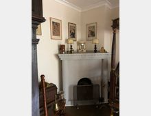 Wohnzimmer mit klassischem Kamin. Der Kamin hat eine weiße Umrandung und ist mit einer Uhr, Lampen und kleinen Deko-Objekten geschmückt; darüber hängen gerahmte Bilder an einer beigen Wand. Im Vordergrund sind antike Möbel und historische Dekorationsgegenstände sichtbar.