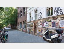 Straßenszene mit einem Gebäude aus roten Ziegeln und weißem Putz, dessen Fassade mit Graffiti und Plakaten bedeckt ist. Daneben verläuft ein gepflasterter Gehweg, gesäumt von geparkten Fahrrädern und einem weißen Motorroller, mit Bäumen im Hintergrund.