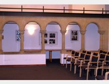 Innenraum mit historischen Steinarkaden und modernen Sitzstühlen. Im Hintergrund sind eine weiße Wand mit Bildern und eine angestrahlte Leuchte sichtbar.