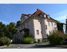 Zweistöckige Villa mit hellem Putz und rotem Walmdach, flankiert von großen Säulen am Eingangsbereich. Das Gebäude liegt in einer grünen Umgebung mit Büschen und Bäumen, Fahrräder stehen vor der Fassade.