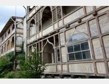 Holzgebäude mit verzierten Spitzbögen und Gitterelementen an den Fassaden, teilweise verfallen und von Vegetation umgeben. Die Fassade zeigt verblasste Farben und verwitterte Fenster in einem ländlichen, bewachsenen Umfeld.