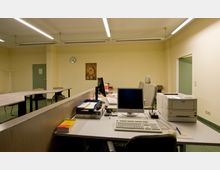 Büro- oder Konferenzraum mit langen Tischen und Stühlen, einer hellgelben Wand, einem Computer-Arbeitsplatz mit Monitor, Tastatur und Drucker im Vordergrund. Im Hintergrund befinden sich ein Poster, eine Wanduhr und eine grüne Tür.