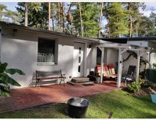 Kleines Gebäude mit weißer Fassade, davor eine Terrasse aus roten Backsteinen. Auf der linken Seite steht eine Metallbank, rechts befindet sich eine überdachte Sitzecke mit Stühlen und Pflanzen. Dahinter sind hohe Nadelbäume sichtbar.