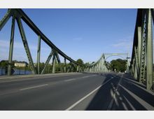 Grüne Stahlbrücke mit geschwungenen Bögen und Gitterträgern über eine Straße, flankiert von Bäumen und einem Gewässer im Hintergrund, unter klarem, blauem Himmel.