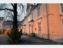 Historisches Gebäude mit heller Fassade und großen Fenstern, flankiert von einem Baum mit kahlen Ästen. Im Vordergrund ein gepflasterter Platz mit Weihnachtsbaum und roten Holzhütten im Hintergrund.