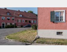 Reihenhäuser mit rosa Fassaden und grünen Fensterläden in einer ruhigen Wohnsiedlung. Im Vordergrund eine Straße, ein kleiner Grünstreifen und ein parkendes Auto, im Hintergrund ein weiteres Haus.