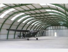 Ein Flugzeughangar mit grüner Stahlkonstruktion und weißen, gewölbten Dachelementen. In der Mitte steht ein kleines, olivgrünes Propellerflugzeug auf einer gepflasterten Fläche.