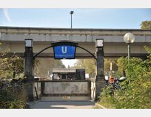 Eingang zur U-Bahn-Station Breitenbachplatz in Berlin, mit einem blauen Schild und einem darüberliegenden Brückensegment. Im Vordergrund befinden sich Lampen, Fahrräder und grüne Büsche, während im Hintergrund ein Imbissstand zu sehen ist.