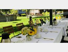 Langer, gedeckter Tisch mit weißen Tischdecken im Freien, dekoriert mit Blumenvasen aus Sonnenblumen und Hortensien. Im Hintergrund grüner Rasen, Holzmöbel und ein Pavillon in einer Gartenumgebung.