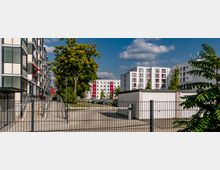 Mehrstöckige Wohngebäude mit weißer Fassade und roten Akzenten stehen hinter einem Tor und einer gepflasterten Fläche. Im Vordergrund sind ein Baum, ein Zaun und ein kleiner Flachbau sichtbar.