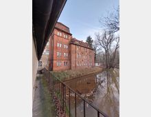 Backsteinbau am Ufer eines ruhigen Gewässers, umgeben von kahlen Bäumen. Ein schmiedeeisernes Geländer verläuft entlang eines schmalen Weges neben dem Gebäude.