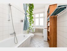 Helles, gefliestes Badezimmer mit Badewanne und Duscharmatur an der linken Wand sowie einem großen Fenster über einem Heizkörper am Ende des Raums. Rechts steht ein Holzregal mit Handtüchern neben einer Waschmaschine; eine hängende Grünpflanze und ein gemusterter Duschvorhang setzen Akzente.