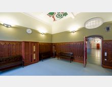 Raum mit Holzwandverkleidungen und schlanken gotischen Verzierungen, mit einer Uhr an einer grün gestrichenen Wand. Es gibt Holzbänke, eine Tür mit Rundbogen führt zu einem angrenzenden Bereich mit dekorativen Wandmalereien.