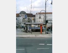 Ansicht des U-Bahnhofs Hermannplatz in Berlin, mit einem Stationsschild und einem Eingang, umgeben von städtischer Architektur mit mehrstöckigen Gebäuden im historischen Stil. Im Vordergrund befinden sich eine Straße, Fahrradständer und kahle Bäume, während eine goldene Statue prominent auf einem Sockel über dem Eingang steht.